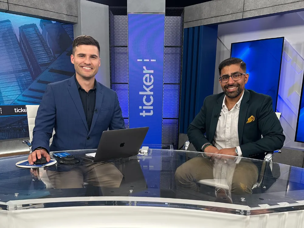 Two professional men sit at a desk in a modern blue-lit studio with a laptop between them, smiling at the camera.